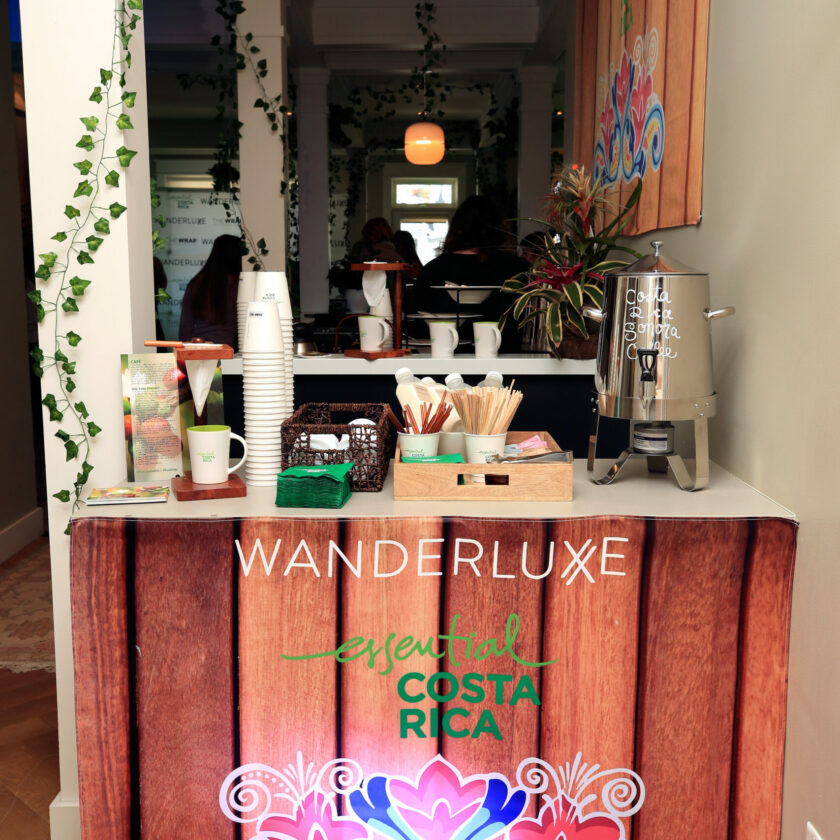 A cozy coffee station at an event branded with 'Wanderluxe, Essential Costa Rica' featuring a pour-over coffee setup, with informational pamphlets and a vibrant display of indoor plants enhancing the tropical theme.