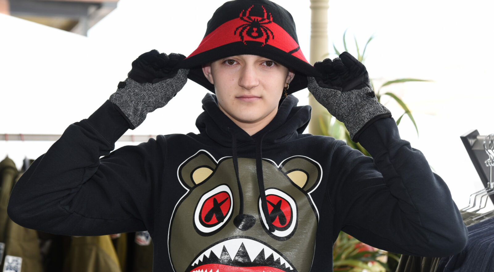 A young man adjusting a red and black hat on his head while wearing a black hoodie with a cartoon bear design.