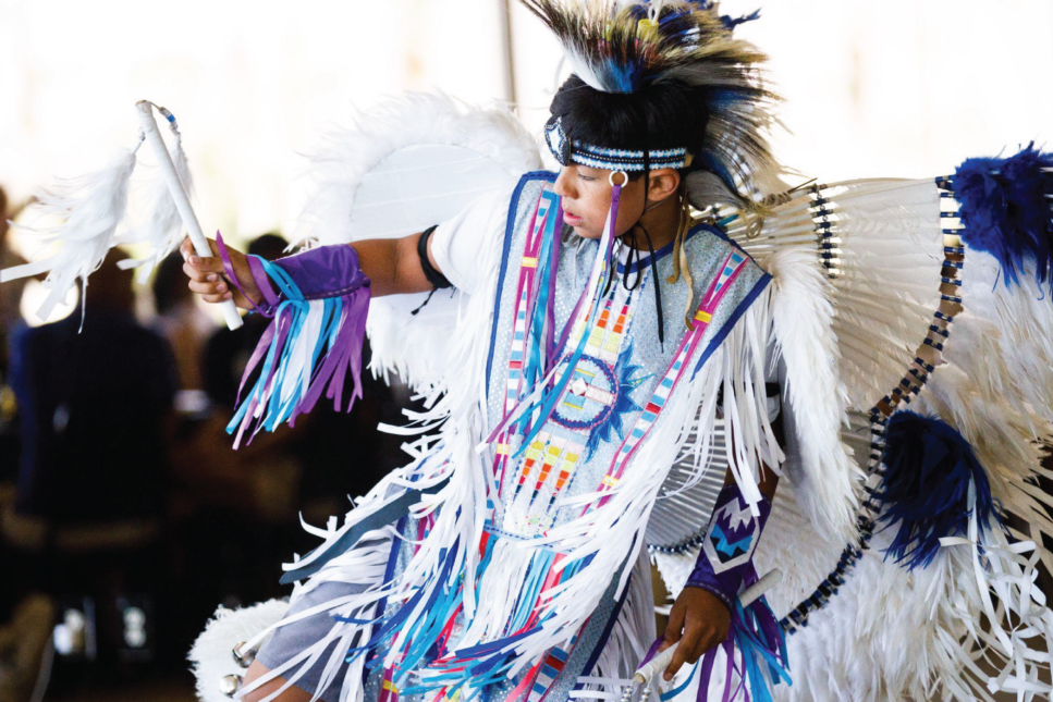 Native American dancer in traditional ceremonial attire with a feathered headdress and colorful ribbons.