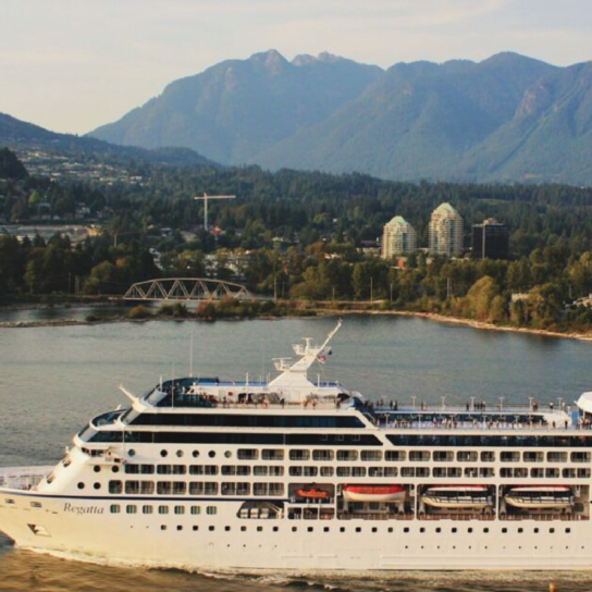 The cruise ship Oceania Regatta glides through calm waters near Vancouver, Canada, framed by lush green hills and the urban skyline in the background. This scenic vista highlights the vessel's sleek design and bustling decks as passengers enjoy the panoramic views.