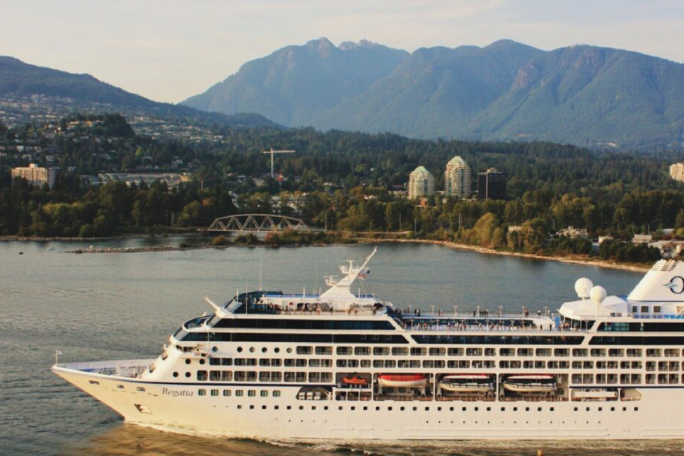 The cruise ship Oceania Regatta glides through calm waters near Vancouver, Canada, framed by lush green hills and the urban skyline in the background. This scenic vista highlights the vessel's sleek design and bustling decks as passengers enjoy the panoramic views.