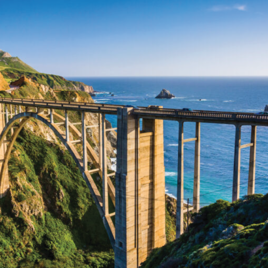 Breathtaking view of the iconic Bixby Bridge along California's Highway 1, framed by lush greenery and overlooking the vivid blue Pacific Ocean.