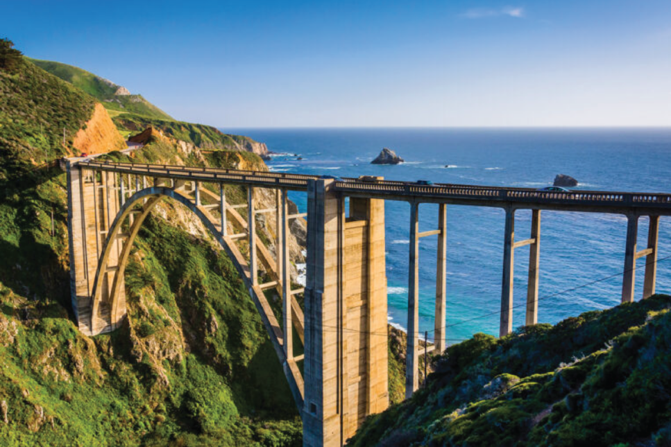 Breathtaking view of the iconic Bixby Bridge along California's Highway 1, framed by lush greenery and overlooking the vivid blue Pacific Ocean.