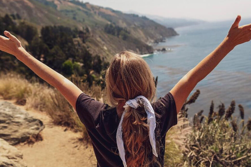 Woman with arms raised in joy overlooking the scenic coastline from a cliff on California's Highway 1.