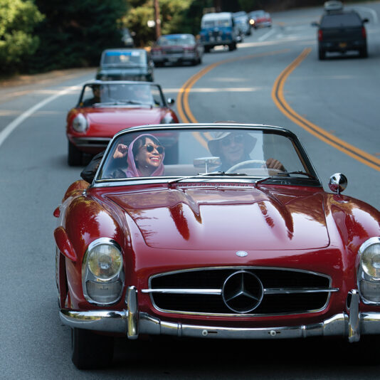 Joyful couple enjoying a scenic drive along a road in a classic red Mercedes convertible, part of a caravan of vintage cars.