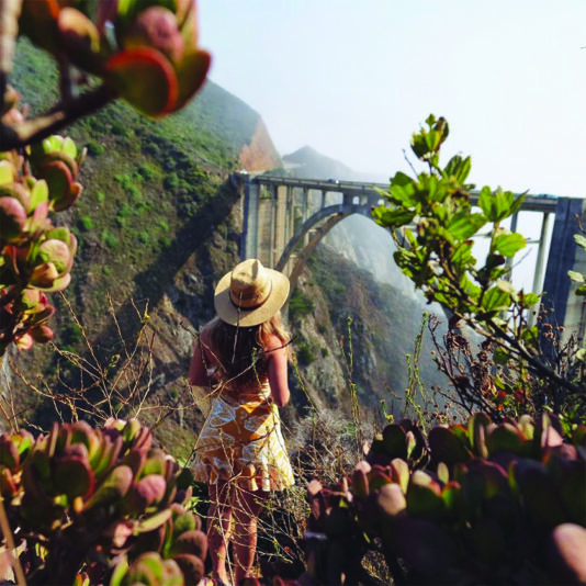 Woman observing the iconic Bixby Bridge amid lush surroundings, embodying the spirit of exploration along California's Highway 1.