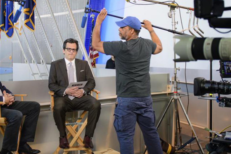 Behind-the-scenes at a news studio, featuring a well-dressed news anchor seated with a tablet in front of cameras, while a crew member adjusts lighting equipment.